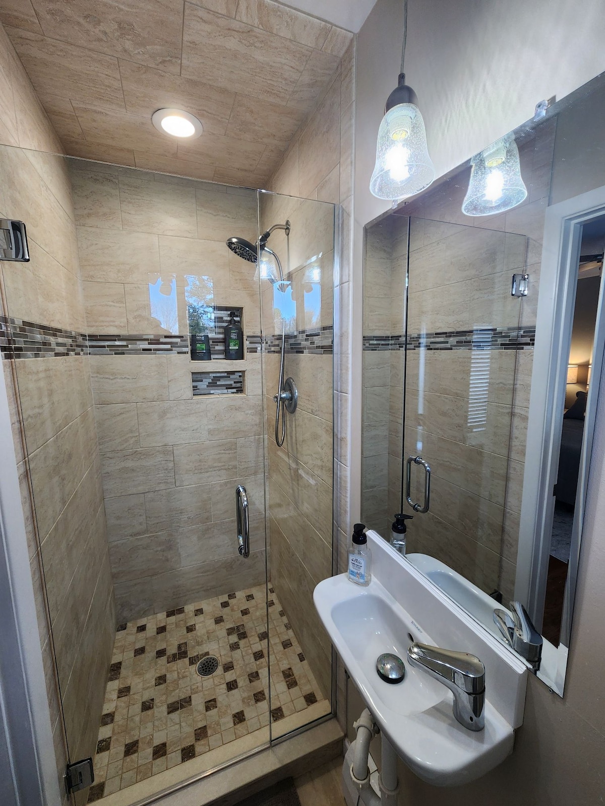 A modern shower is enclosed in glass, featuring rainfall and handheld showerheads. Textured tiles cover the walls and floor, complemented by a small, sleek sink equipped with a polished faucet. Bright lighting is provided by stylish pendant fixtures above.