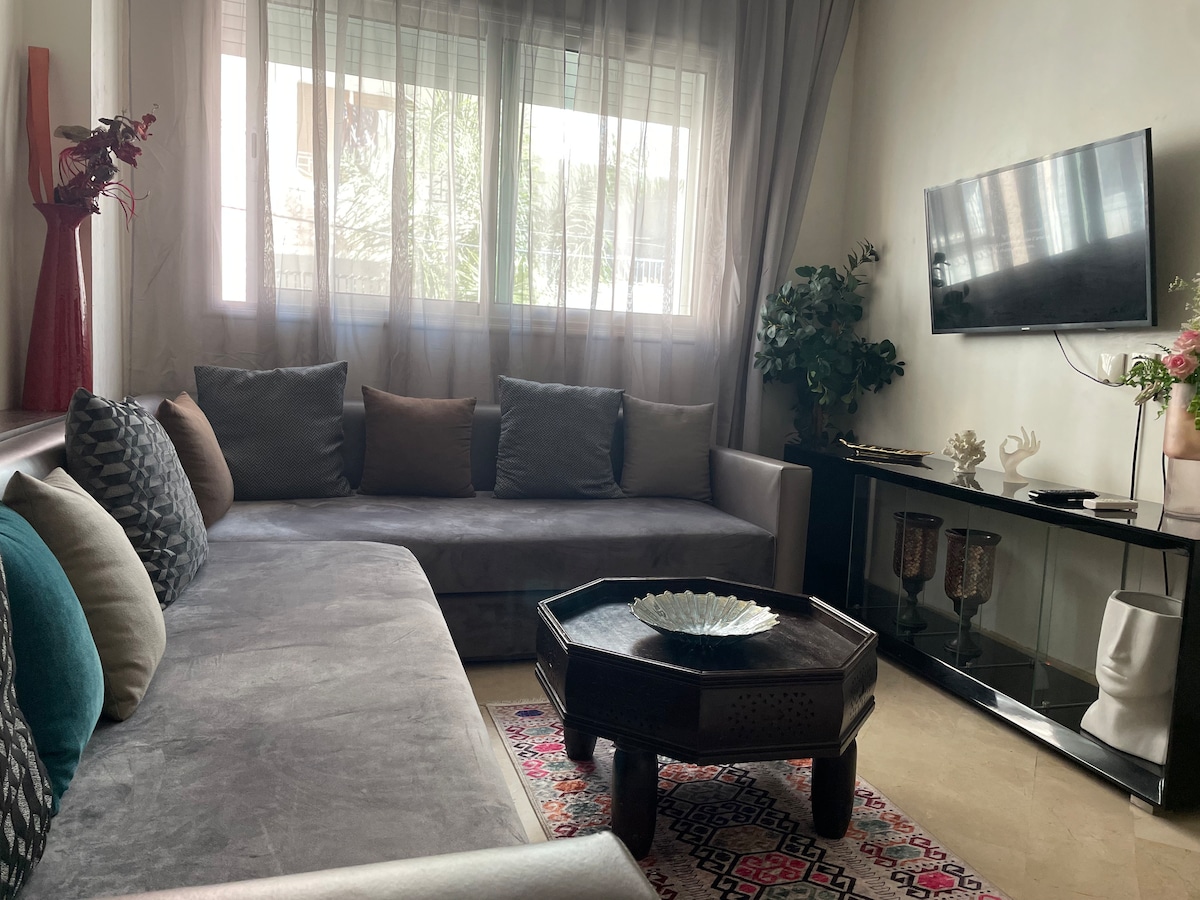 A contemporary living area features a spacious gray sectional sofa with colorful decorative pillows. A black console table holds decorative items and a vase. Natural light filters through sheer curtains, while a flat-screen TV is mounted on the wall, enhancing the neutral and inviting decor.
