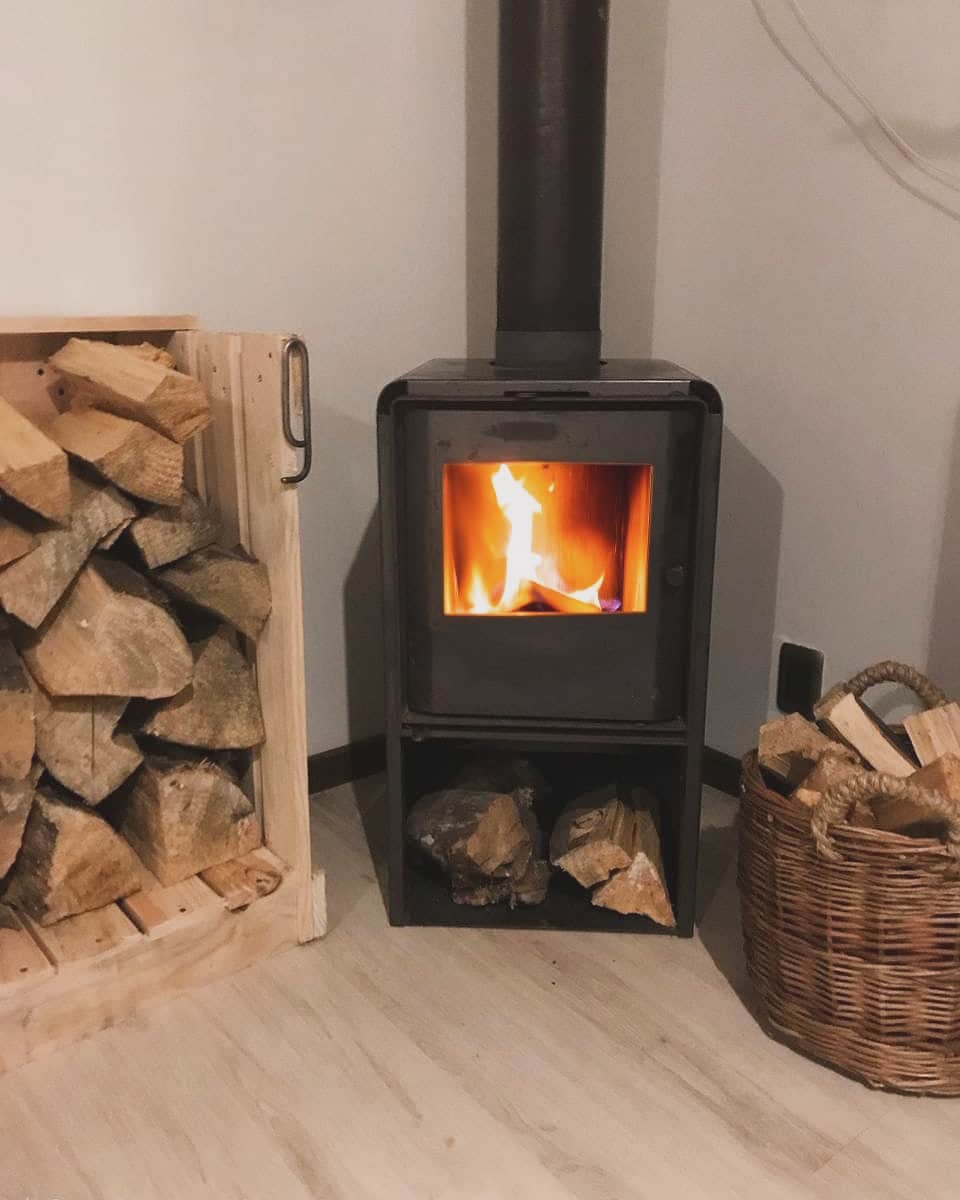 A wood-burning stove is positioned in a corner, with flames visible through the glass door. A stack of firewood is neatly arranged in a wooden holder beside the stove, while additional logs are stored in a woven basket nearby.