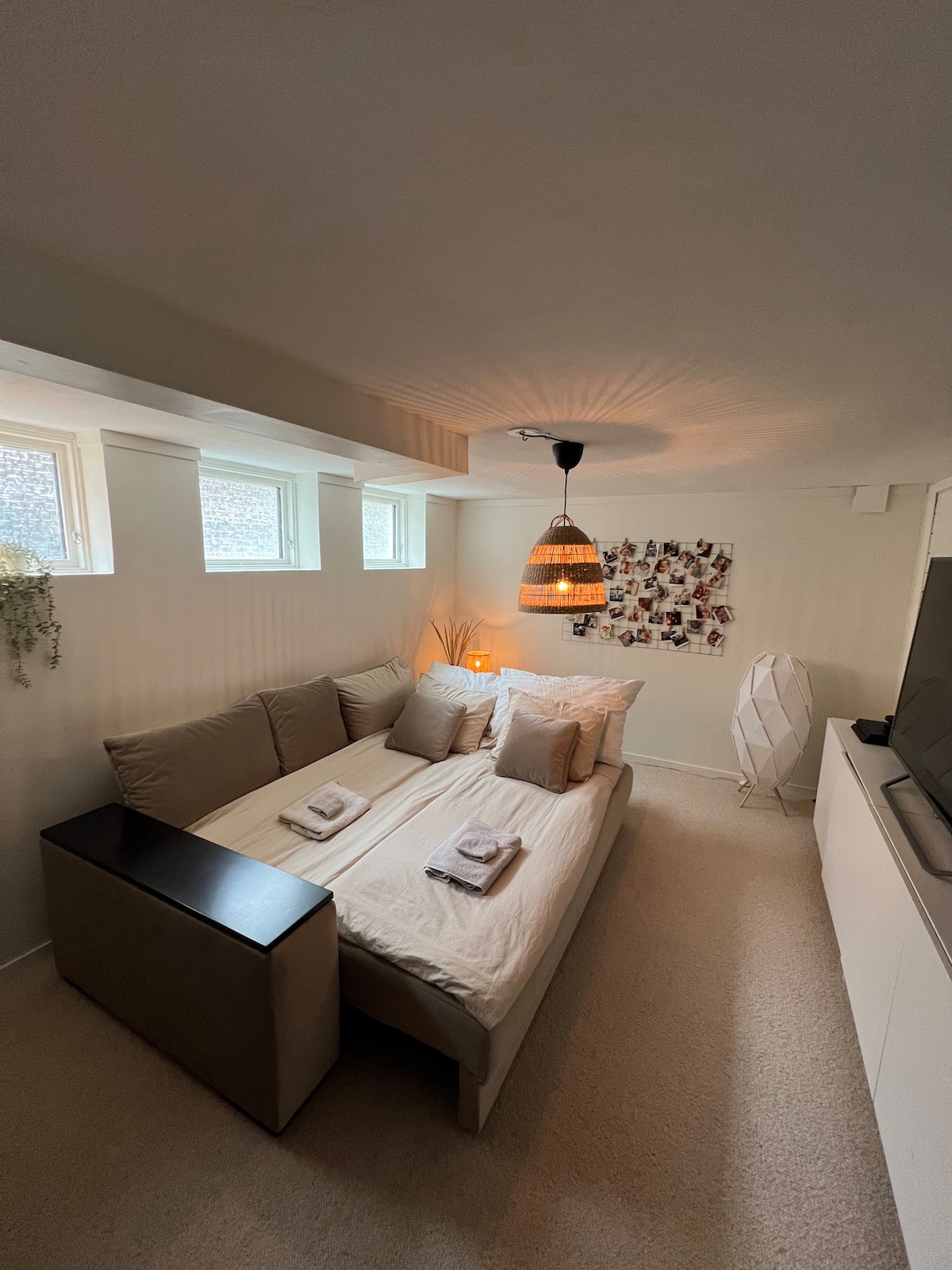 A cozy sleeping area features a spacious bed adorned with neatly arranged towels. Soft lighting is provided by a pendant lamp, while two windows allow natural light to brighten the room. A decorative wall displays an arrangement of photographs, enhancing the inviting ambiance.