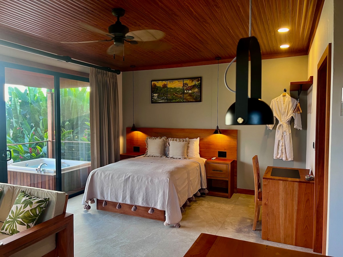 A spacious bedroom is presented, featuring a large bed with neutral-toned bedding. A small desk and cozy chair are positioned nearby. A sliding glass door leads to a balcony with views of lush greenery. A jacuzzi is visible in the corner, enhancing the relaxing ambiance.