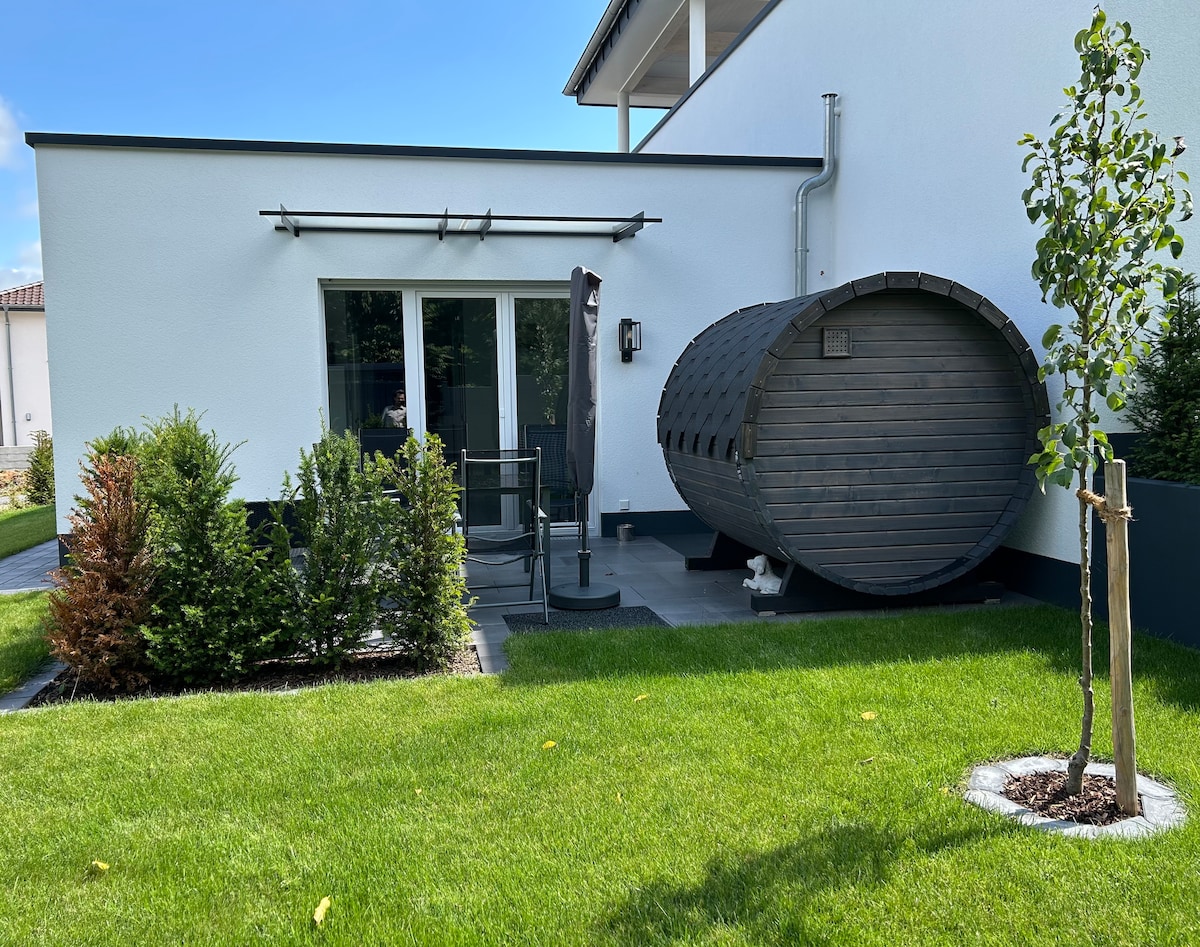 An outdoor area is presented with manicured green grass, flanked by neatly trimmed shrubs. A wooden barrel-shaped sauna is positioned on the right, accompanied by a small seating arrangement consisting of a table and chairs set against the exterior wall of the house.
