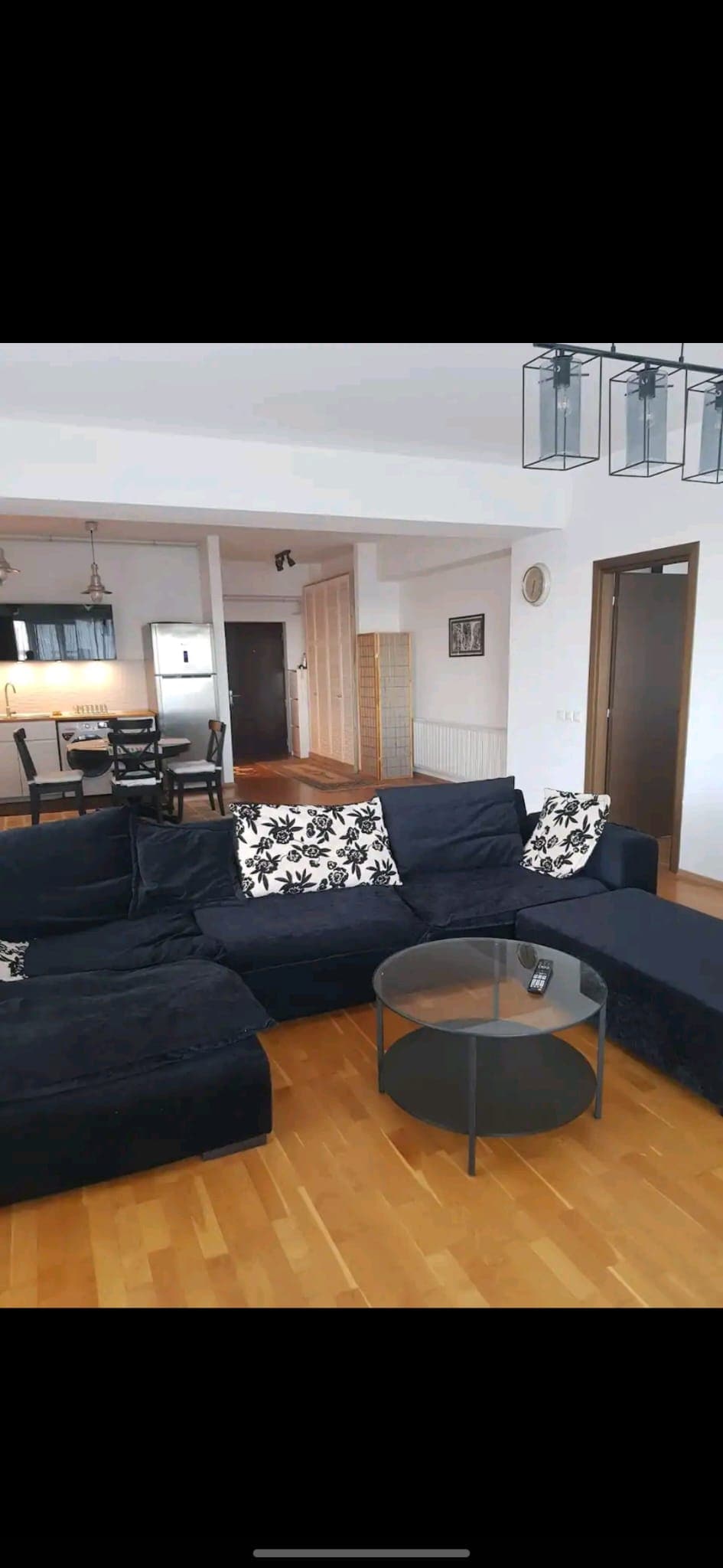 A spacious living area features a dark sectional sofa adorned with decorative cushions. A circular glass coffee table sits in front, complementing the hardwood flooring. The dining area and kitchen are visible in the background, with elegant lighting fixtures hanging above.
