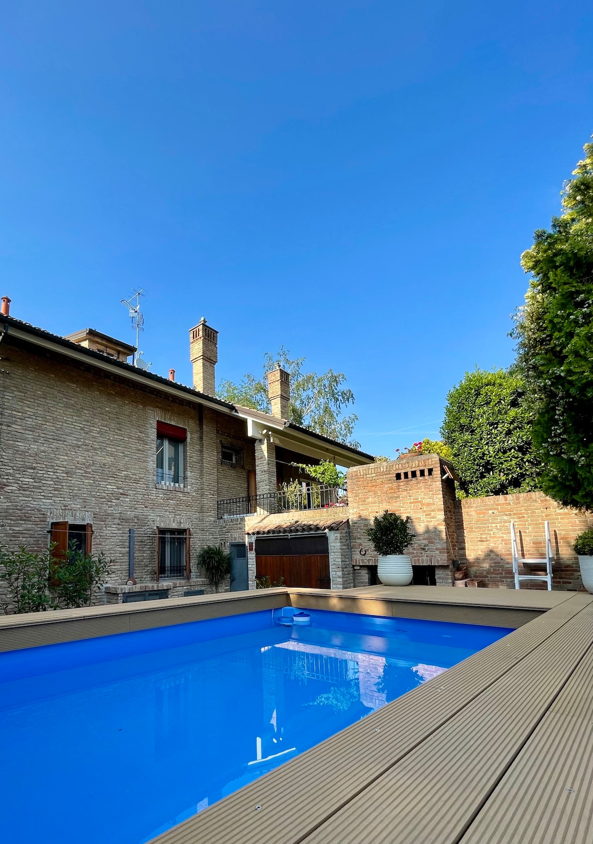 The image shows a private swimming pool surrounded by wooden decking. A stone villa with multiple windows is visible in the background, along with lush greenery that provides shade and privacy. The clear blue sky enhances the inviting atmosphere of the outdoor area.