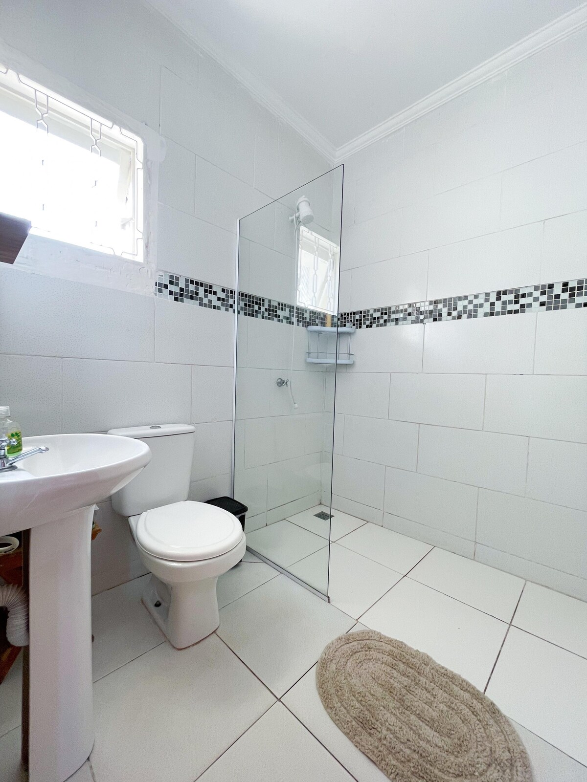 A clean and modern bathroom is displayed, featuring a glass shower enclosure and a compact sink. The walls are tiled in light tones, with a decorative border of black and silver tiles. A round bath mat is positioned in front of the sink.