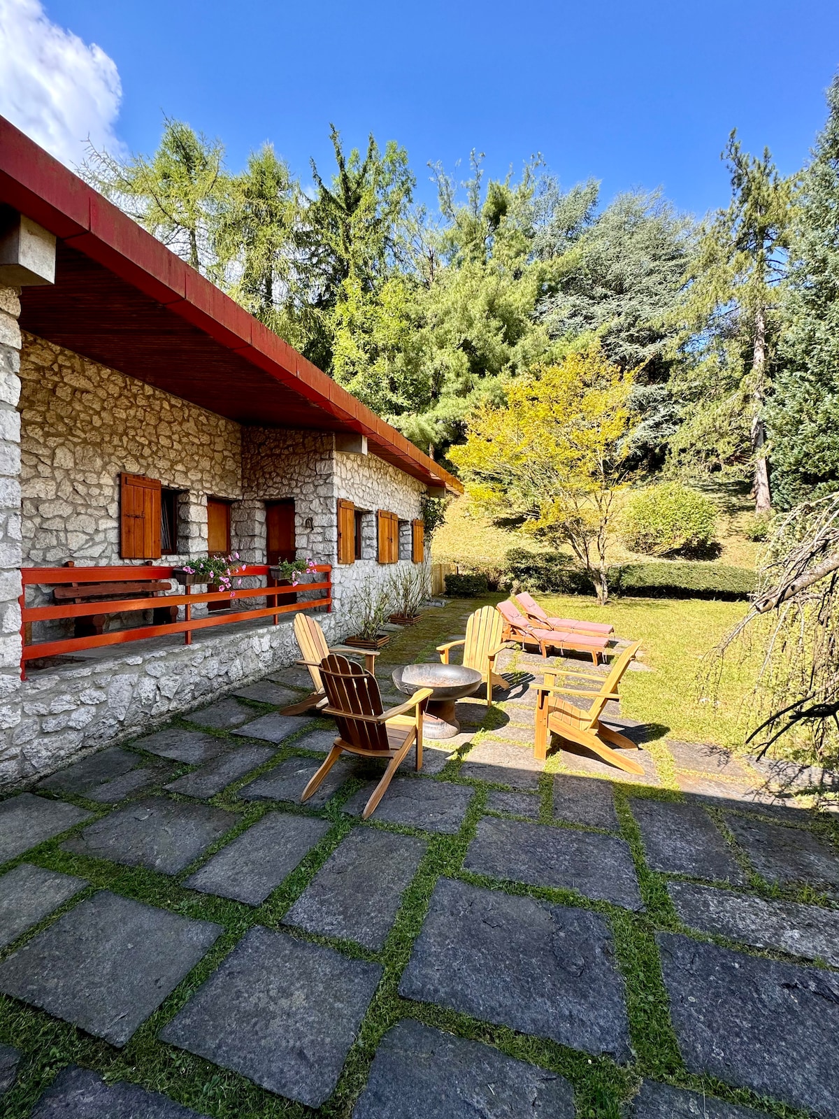 An outdoor seating area is featured, with wooden chairs arranged around a small table on a stone patio. Lush greenery surrounds the space, complemented by sunlight filtering through the trees. The chalet's stone exterior with wooden window shutters is visible in the background.