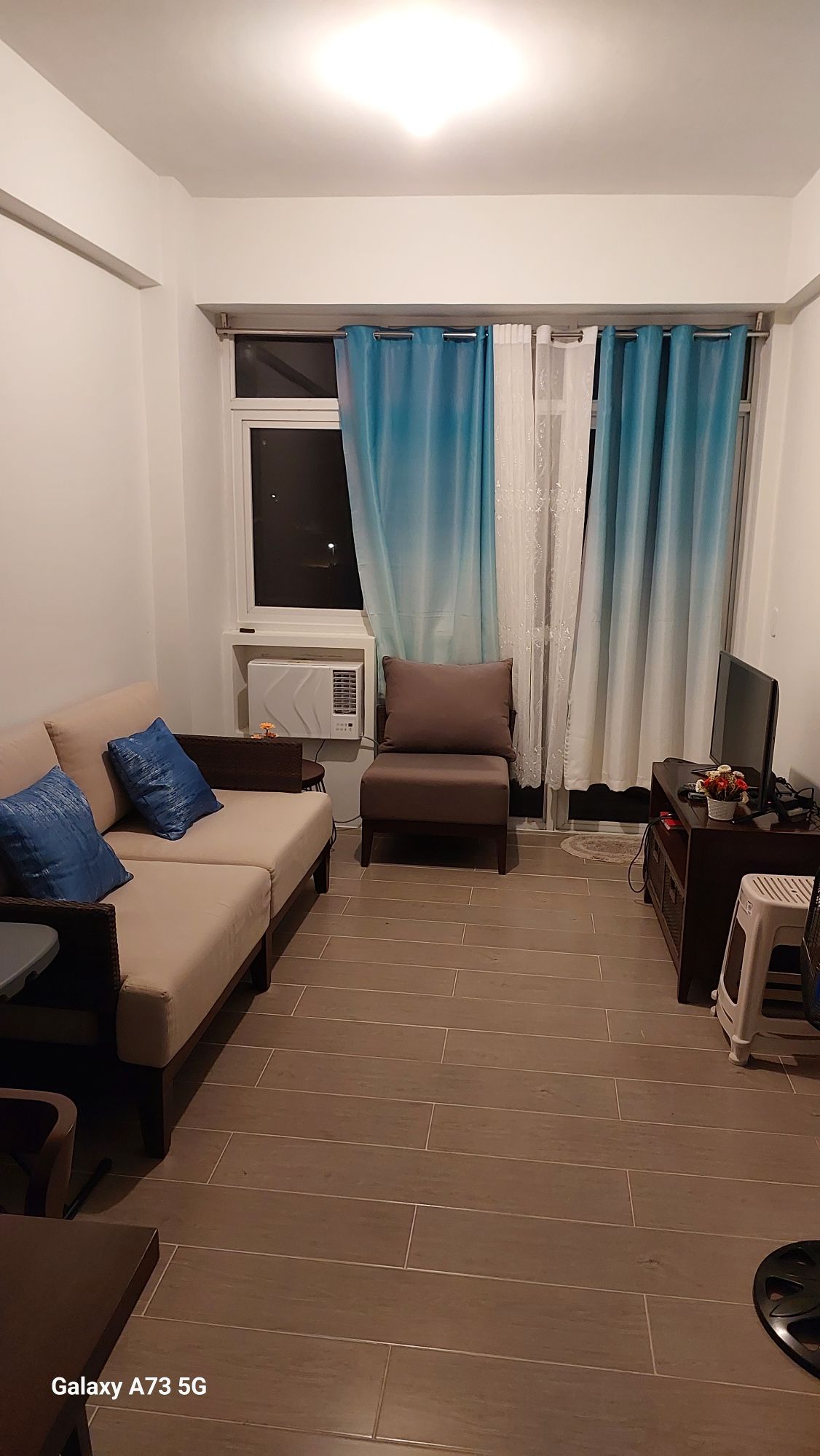 A small living area is presented, featuring a light-colored sofa with blue cushions and a separate armchair. A television is positioned on a dark wood media console, while curtains in an ombre pattern frame a window. Air conditioning is visible in the corner.