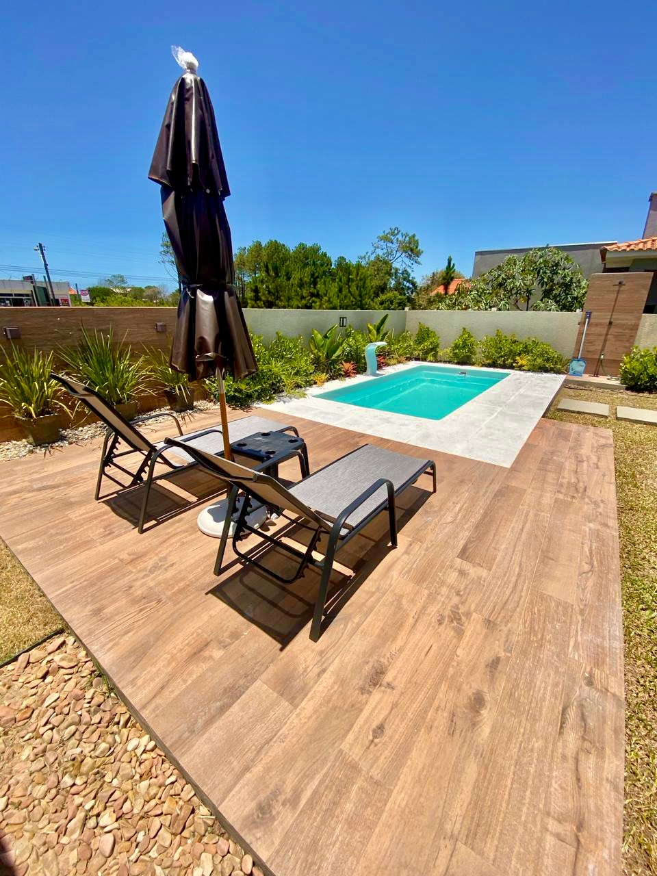 An outdoor space features a private pool surrounded by landscaped greenery. Two lounge chairs are positioned on a wooden deck, accompanied by a large parasol providing shade. The area is designed for relaxation, with a clear blue sky visible above.