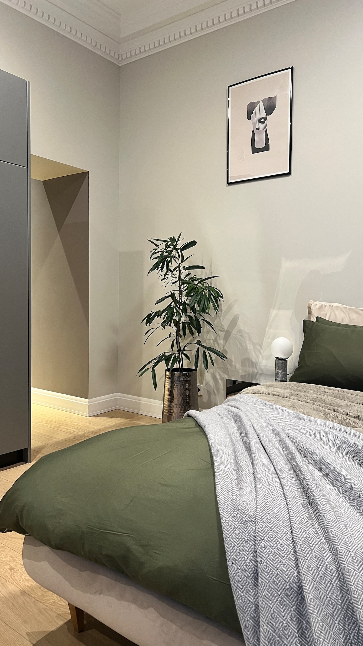A serene bedroom corner is depicted, featuring a spacious bed covered with olive green bedding. A light gray throw is draped casually over the bed. A potted plant adds a touch of greenery, while a framed artwork adorns the wall above, completing the inviting space.
