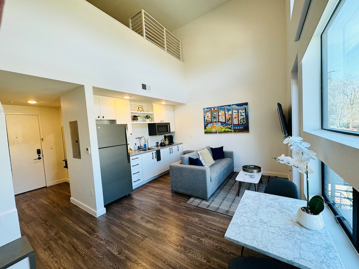 A spacious living area features sleek wooden flooring and high ceilings, creating an open ambiance. A comfortable gray couch is positioned across from a minimalist coffee table. Light streams through large windows, illuminating the modern kitchen and dining area adjacent to the sitting space.