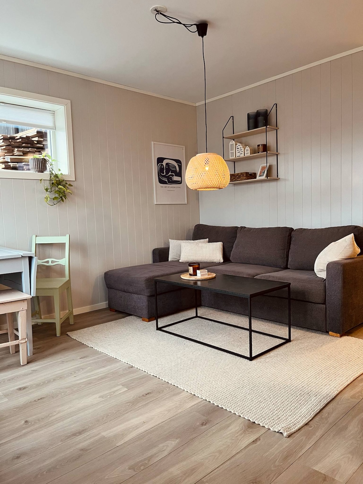A modern living area features a comfortable dark gray sectional sofa accompanied by a minimalist black coffee table. A light fixture hangs above, and a wall shelf displays decorative items. Natural light filters through a window, highlighting the soft beige rug and light wooden flooring.