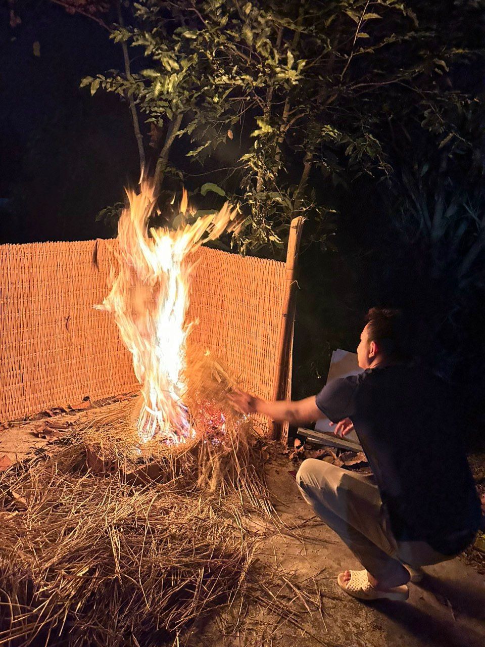 A person is tending to a fire in a natural outdoor setting, surrounded by greenery. The flames rise prominently against the dimly lit background, creating a warm glow. A woven barrier is partially visible in the scene, adding to the rustic atmosphere.