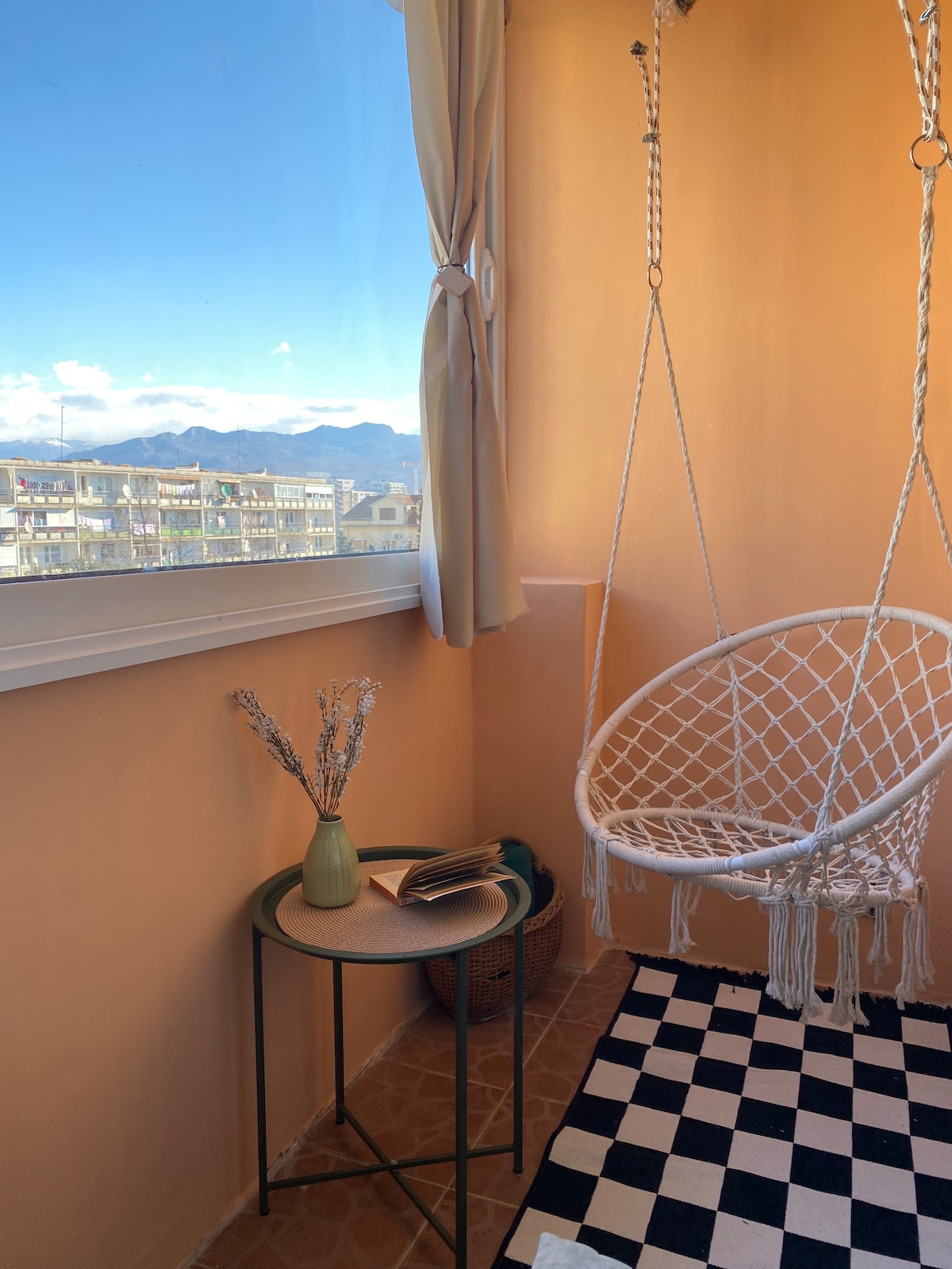 A serene balcony area features a swinging chair and a small table with a vase and a book. Natural light floods in through large windows, revealing distant mountain views. The floor is adorned with a black and white checkered pattern, contributing to the calm ambiance.