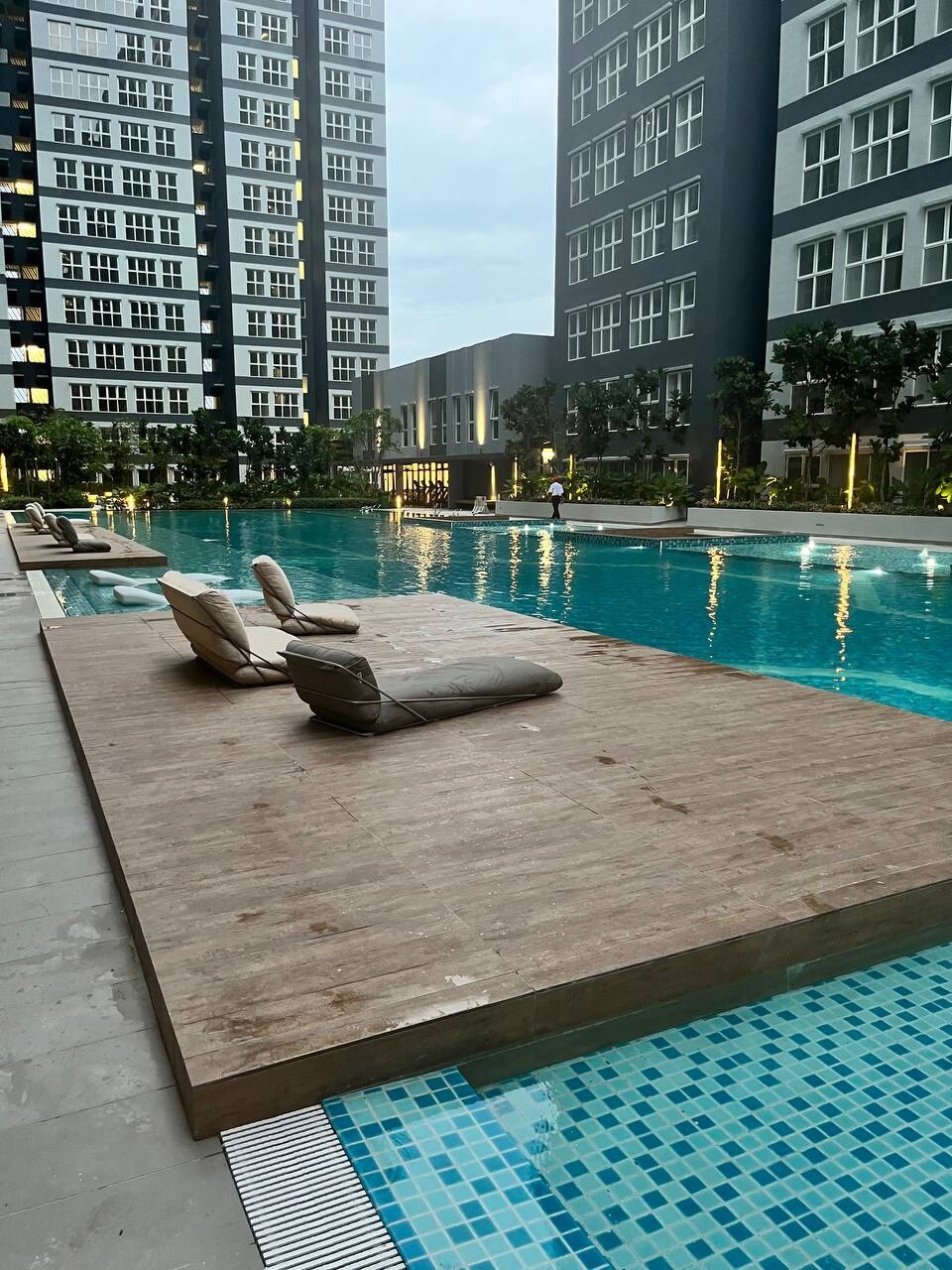 A wooden deck extends over the tranquil pool, featuring two lounge chairs. The pool reflects the surrounding buildings and is bordered by well-manicured greenery. Ambient lighting enhances the serene atmosphere during the evening.