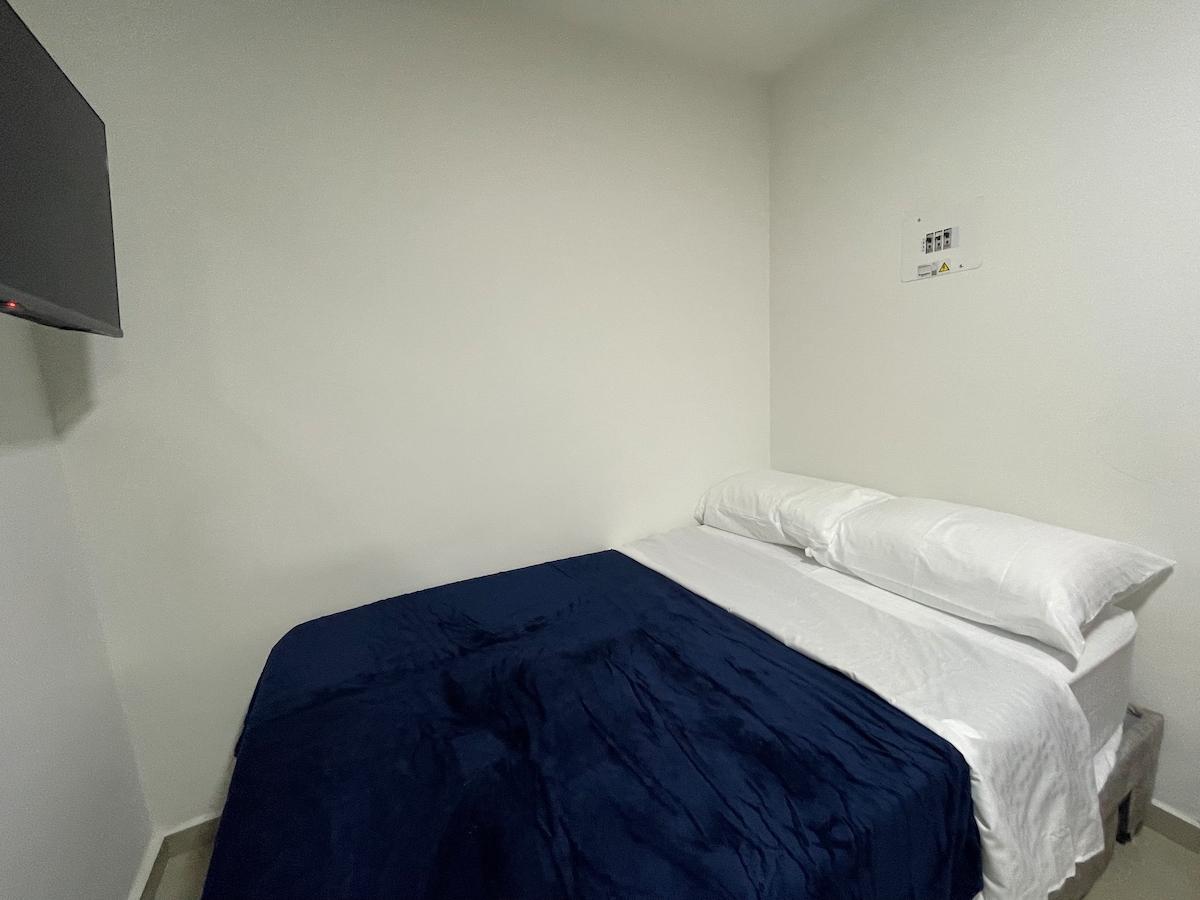 A comfortable sleeping area featuring a spacious bed dressed with a deep blue blanket and crisp white linens. The room is equipped with a mounted television on the wall, and the minimalist decor includes light-colored walls, enhancing the room's simplicity.