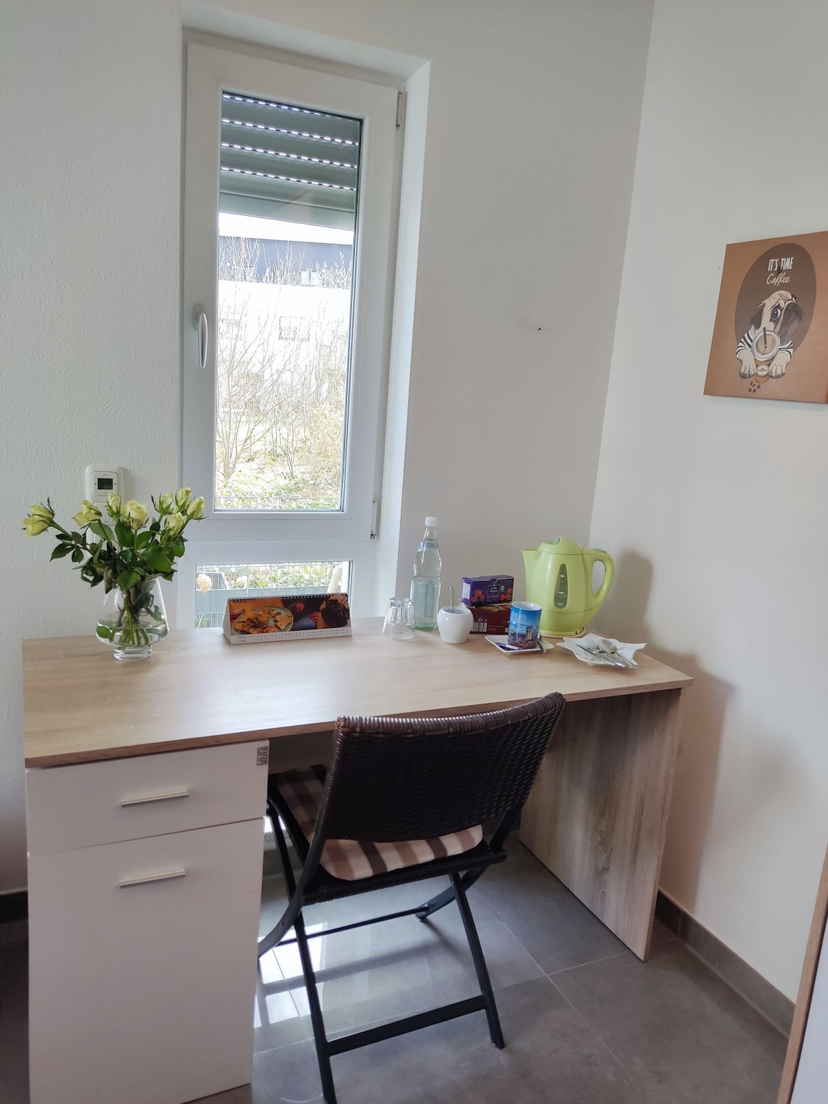A small desk is positioned near a window, with a view of greenery outside. A vase with fresh flowers sits on the desk alongside a kettle, cup, and water bottle. Neutral-colored walls complement the modern design of the space.