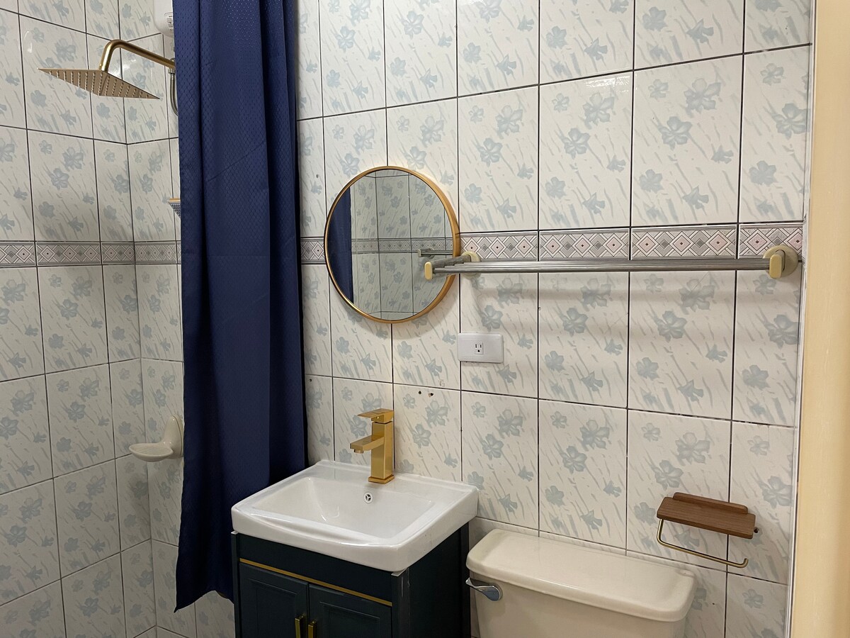 A well-appointed bathroom features tiled walls with a floral pattern. A round mirror is mounted above a sleek vanity with a polished gold faucet. A shower area is enclosed by navy blue curtains, with a showerhead and storage shelf included.