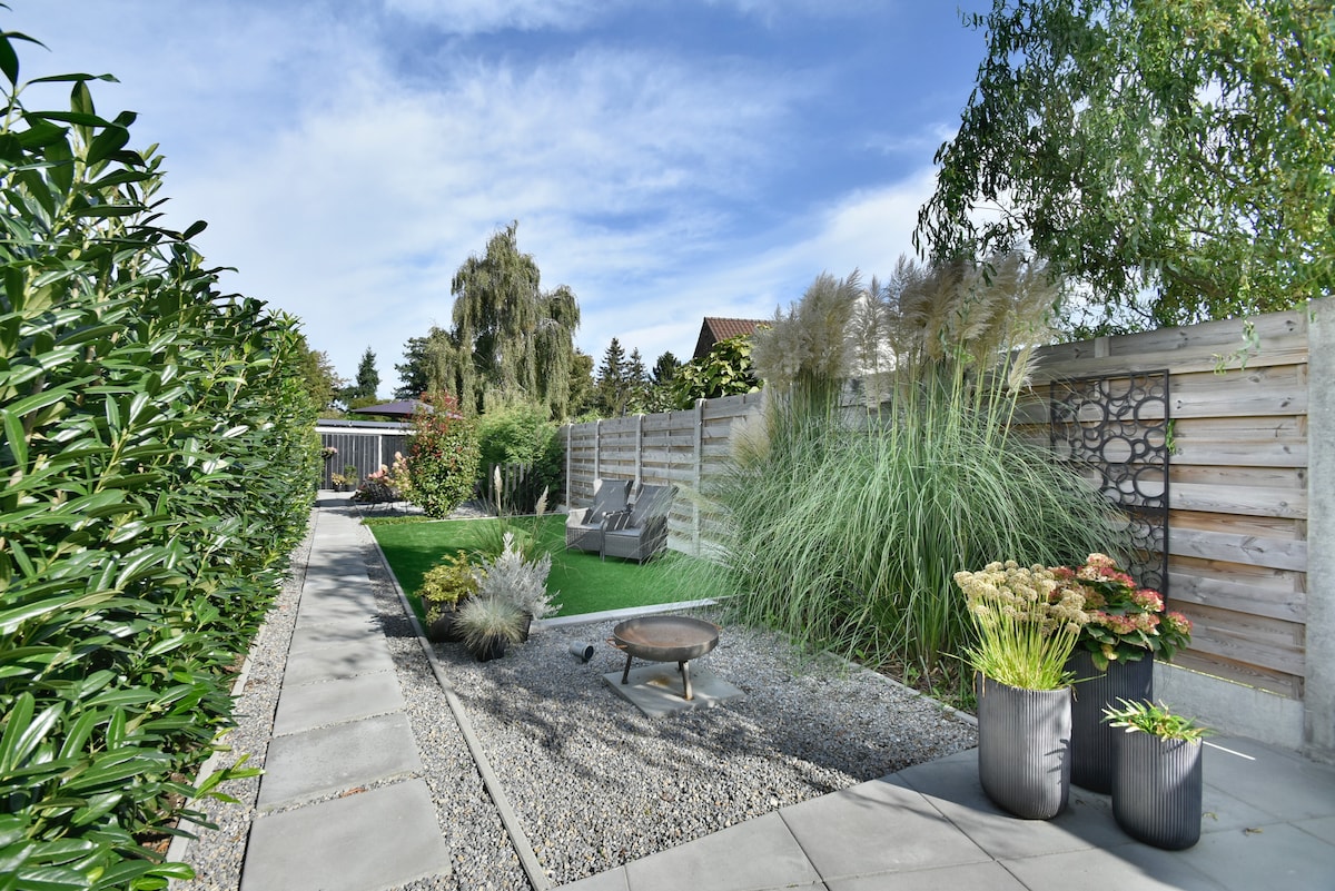 A serene garden area is presented, featuring neatly arranged stone pathways and lush green grass. Tall grasses and seasonal flowers adorn the space, while a fire pit surrounded by pebbles offers a cozy gathering spot. Two lounge chairs are positioned for relaxing outdoors.