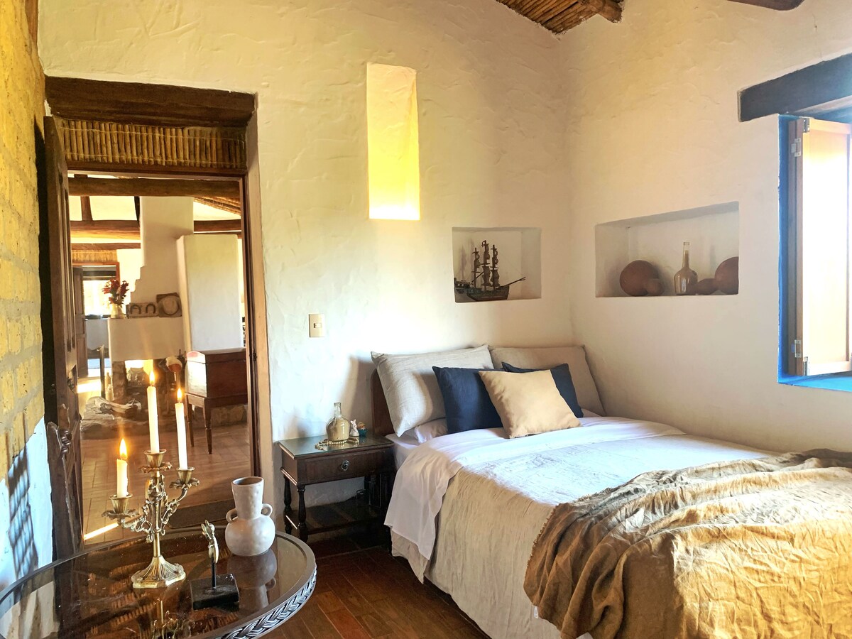A cozy bedroom features a spacious bed with neutral-toned linens, flanked by wooden nightstands. Soft natural light enters through a window, illuminating the room's warm tones. Decorative elements include artwork on the walls and a small table holding candles and a vase.