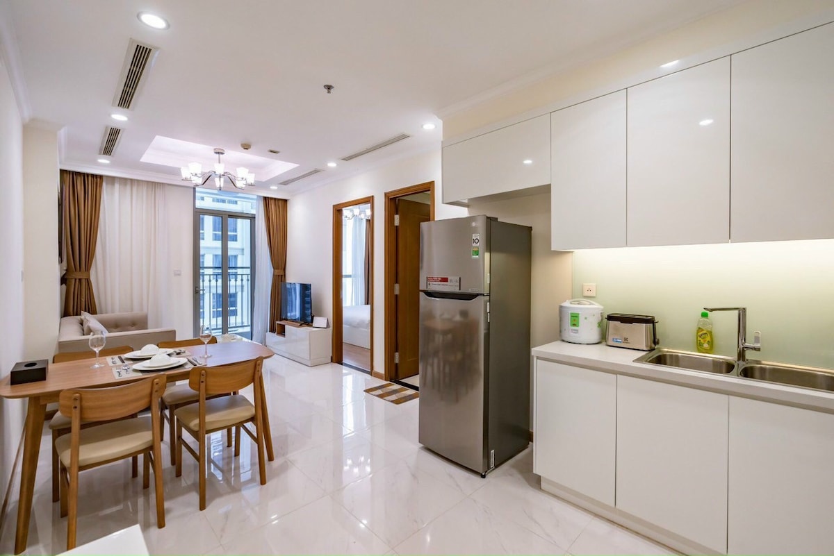 A modern kitchen area features sleek white cabinetry and stainless steel appliances, including a refrigerator. A wooden dining table with four chairs is positioned nearby, enhancing the open concept space. Natural light fills the area through the balcony doors, creating a bright and inviting atmosphere.