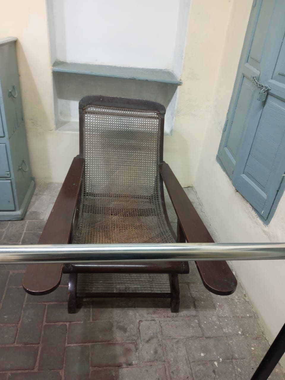 A vintage wooden chair with a woven seat and backrest is positioned against a light-colored wall. Soft natural light filters in through a nearby window, which is framed by blue shutters, creating a calm and inviting corner.