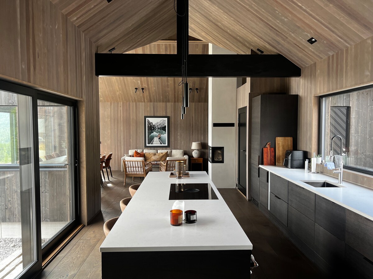 A modern kitchen features a spacious island with a sink and stove, surrounded by wooden cabinetry. Large windows allow natural light to fill the room, which opens to a living area adorned with comfortable seating. High ceilings contribute to an airy atmosphere.