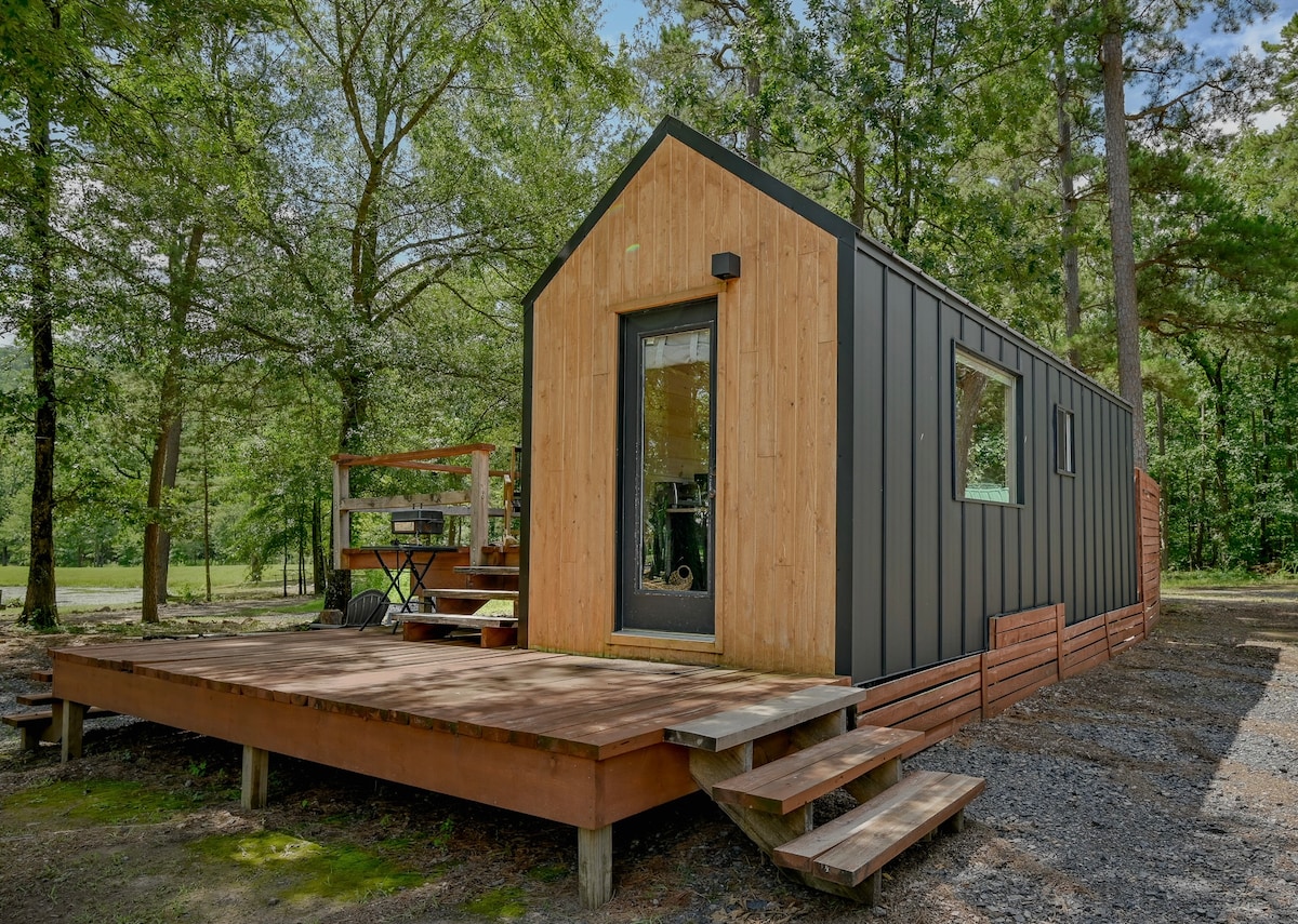 A modern tiny home is set on a wooden deck, surrounded by lush greenery. Large windows allow natural light to illuminate the interior. A cozy outdoor seating area is accessible via steps, creating a welcoming entrance to the tranquil retreat.