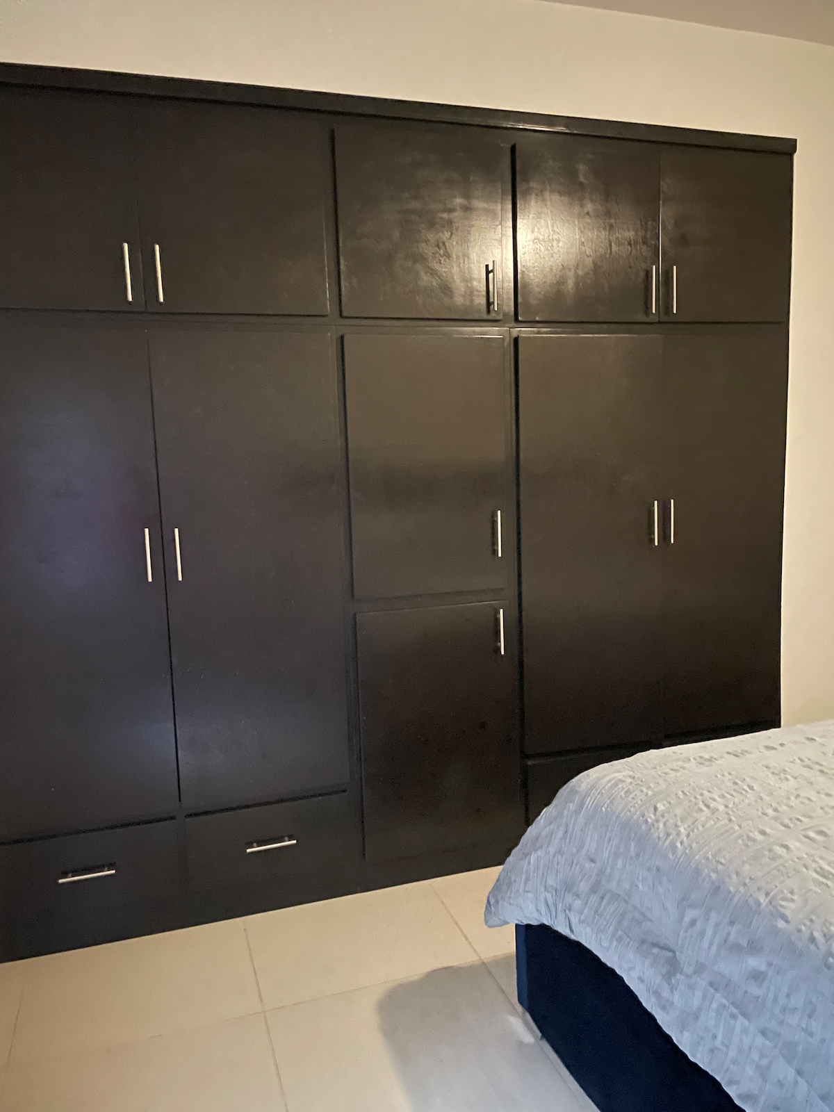 A bedroom is presented featuring a spacious dark wood wardrobe with multiple doors. The wardrobe is complemented by a neatly made bed with a light-colored blanket. Neutral-colored tiles are visible on the floor, contributing to a calm ambiance.
