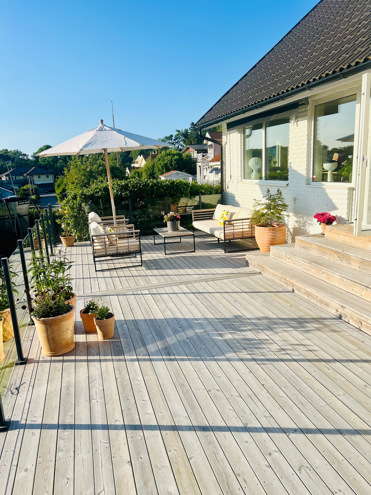 A spacious wooden deck offers a seating area with comfortable chairs and an umbrella for shade. Potted plants are arranged along the railing and steps, enhancing the outdoor space. The view suggests proximity to natural surroundings and provides a welcoming atmosphere.