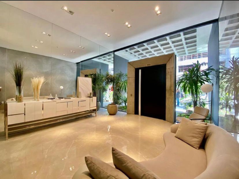 A modern lobby area features sleek surfaces and neutral tones. Lush greenery is positioned throughout, creating a calming environment. A stylish couch invites relaxation, while a glass wall allows natural light to illuminate the space. Two elevators are visible, enhancing accessibility within the building.