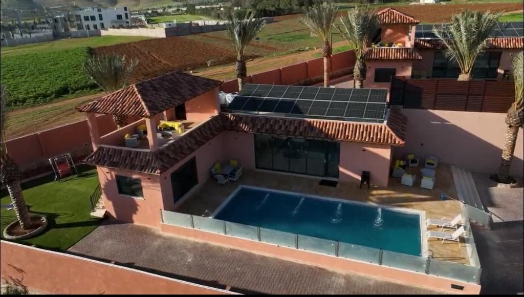 An aerial view presents a pink villa surrounded by palm trees and lush greenery. A private pool is featured in the foreground, complemented by a spacious patio area. Solar panels are visible on the roof, reflecting sustainability in the design.
