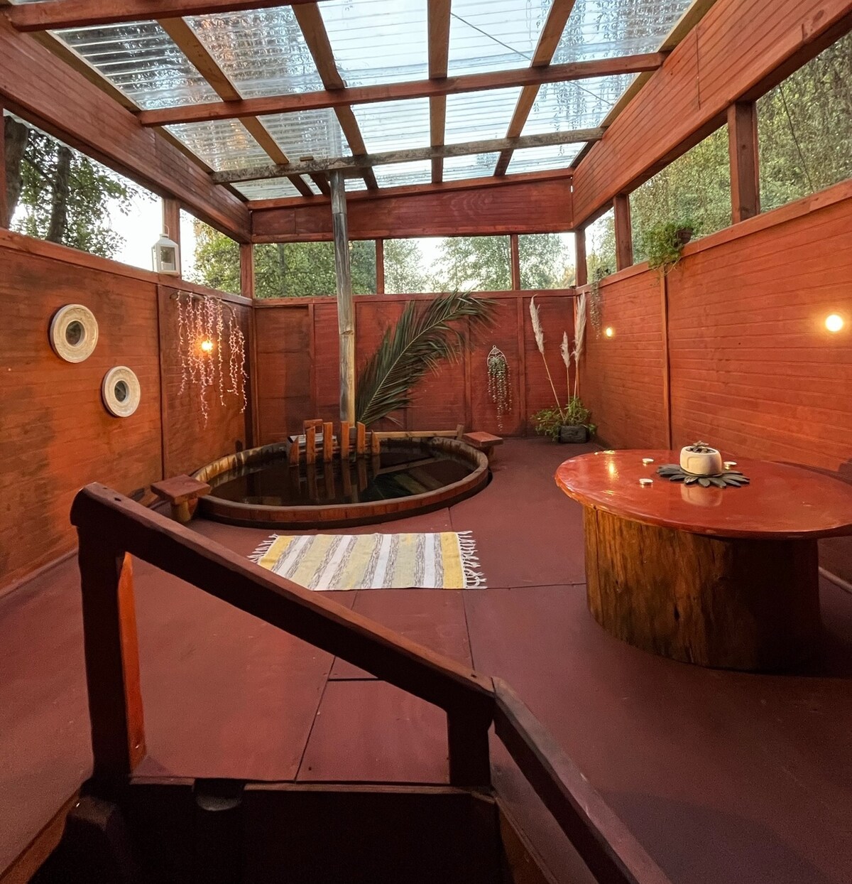 An interior space features wooden walls and a skylight, allowing natural light to enter. A circular tub is situated at the center with a wooden table nearby. Decorative round wall hangings and a patterned rug add visual interest to the serene environment.
