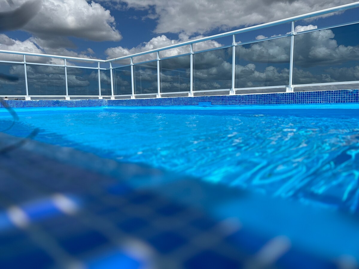 A rooftop pool is shown, featuring clear blue water that reflects the sky. A glass railing surrounds the pool area, providing safety and unobstructed views. Dramatic clouds add depth to the scene, enhancing the serene atmosphere.