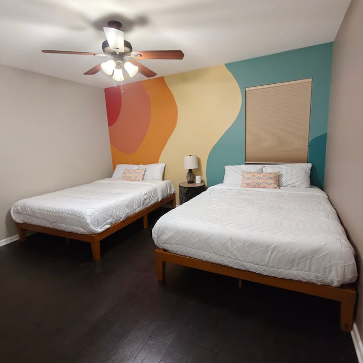 A comfortable bedroom with two queen beds, soft lighting, and natural light from the window. Clean lines and custom, hand painted mural create a restful space suited for shared stays, relaxed evenings, and a good night’s sleep.