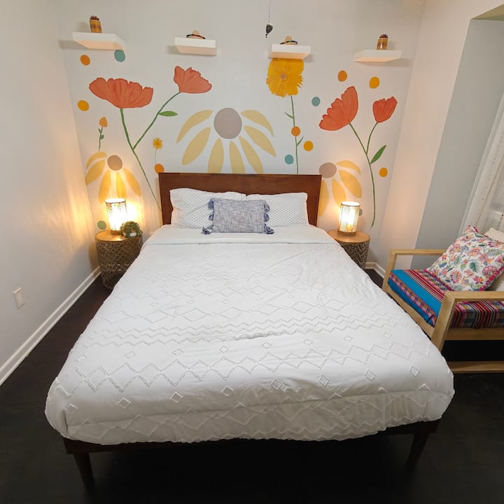 A serene bedroom with a comfortable bed, hand painted mural, and natural light from the window. Thoughtful décor and a cozy seating nook create a calm space for rest, reading, and quiet moments during your stay.