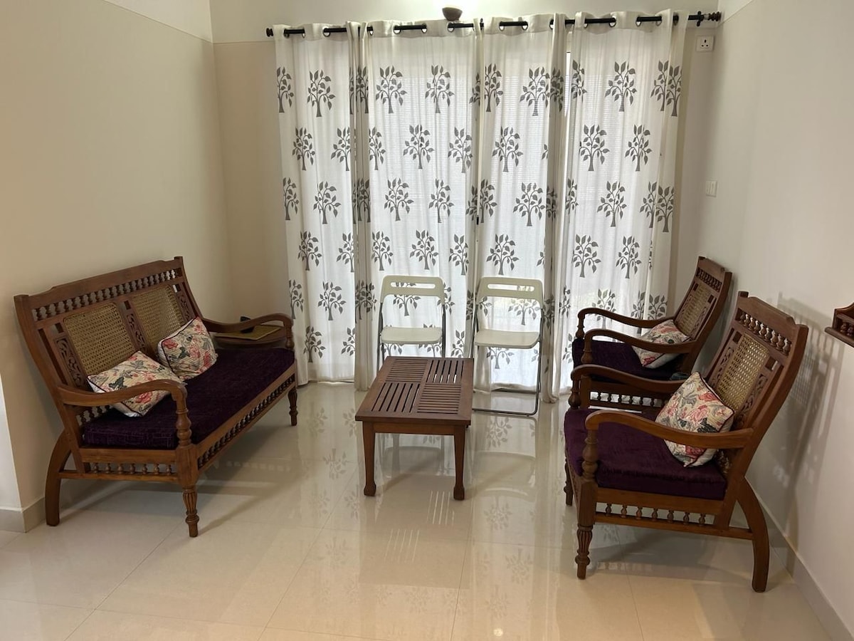 A spacious seating area is furnished with traditional wooden sofas adorned with cushions, accompanied by two simple chairs and a low wooden table. Light filters through sheer curtains embellished with a floral pattern, enhancing the bright and airy ambiance.