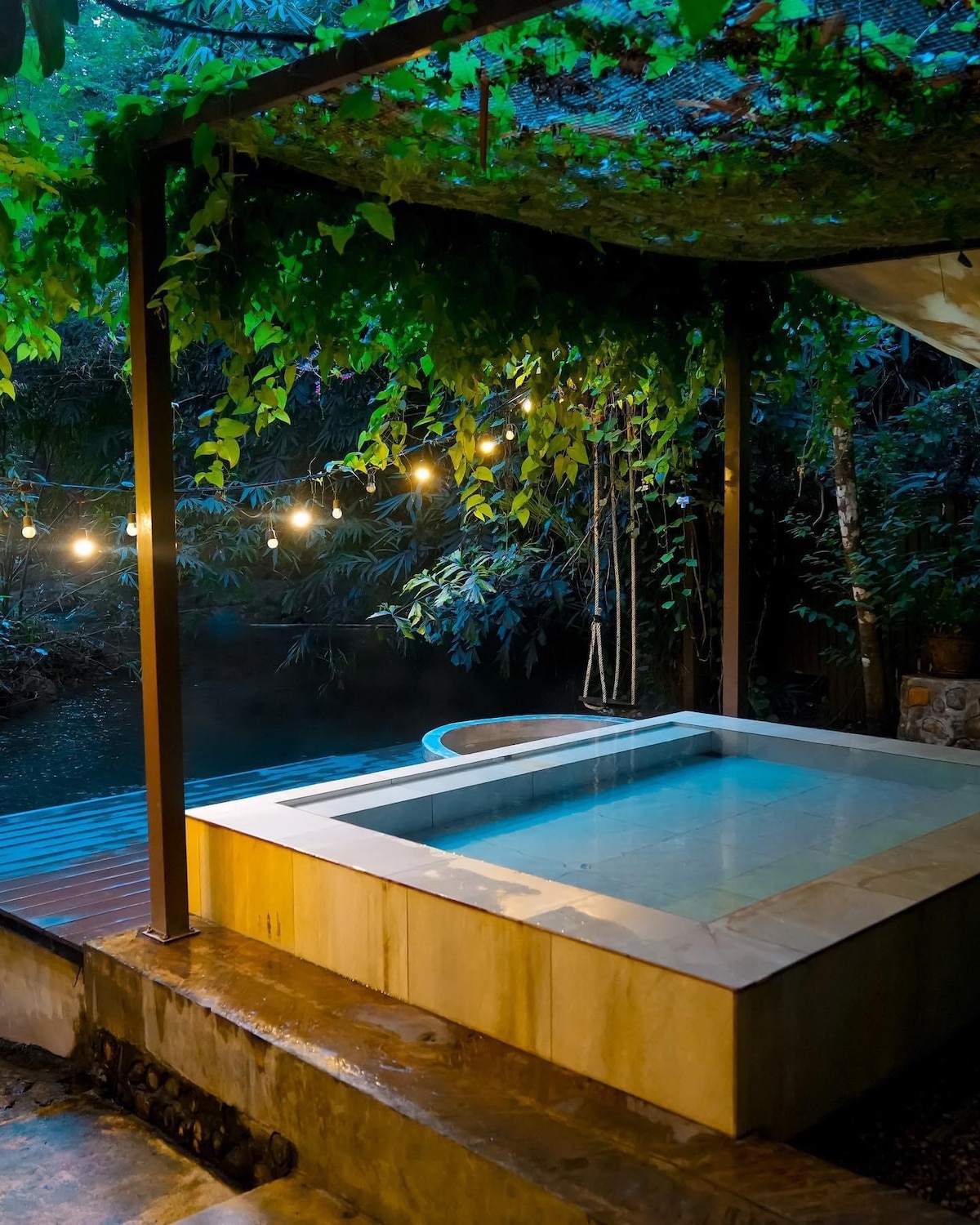 An outdoor hot spring is surrounded by lush greenery, with soft lighting from hanging bulbs creating a serene ambiance. The water in the spa reflects a calm blue hue, set against the tranquil backdrop of the forest.