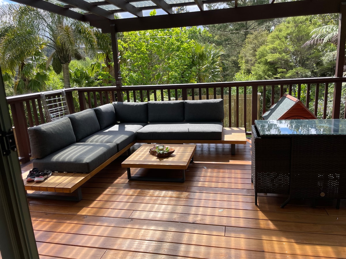 A spacious deck features a comfortable gray sectional sofa accompanied by a low wooden table. Surrounded by lush greenery, this area provides a peaceful spot for relaxation, with sunlight filtering through the overhead trellis. A small storage unit is also visible in the corner.