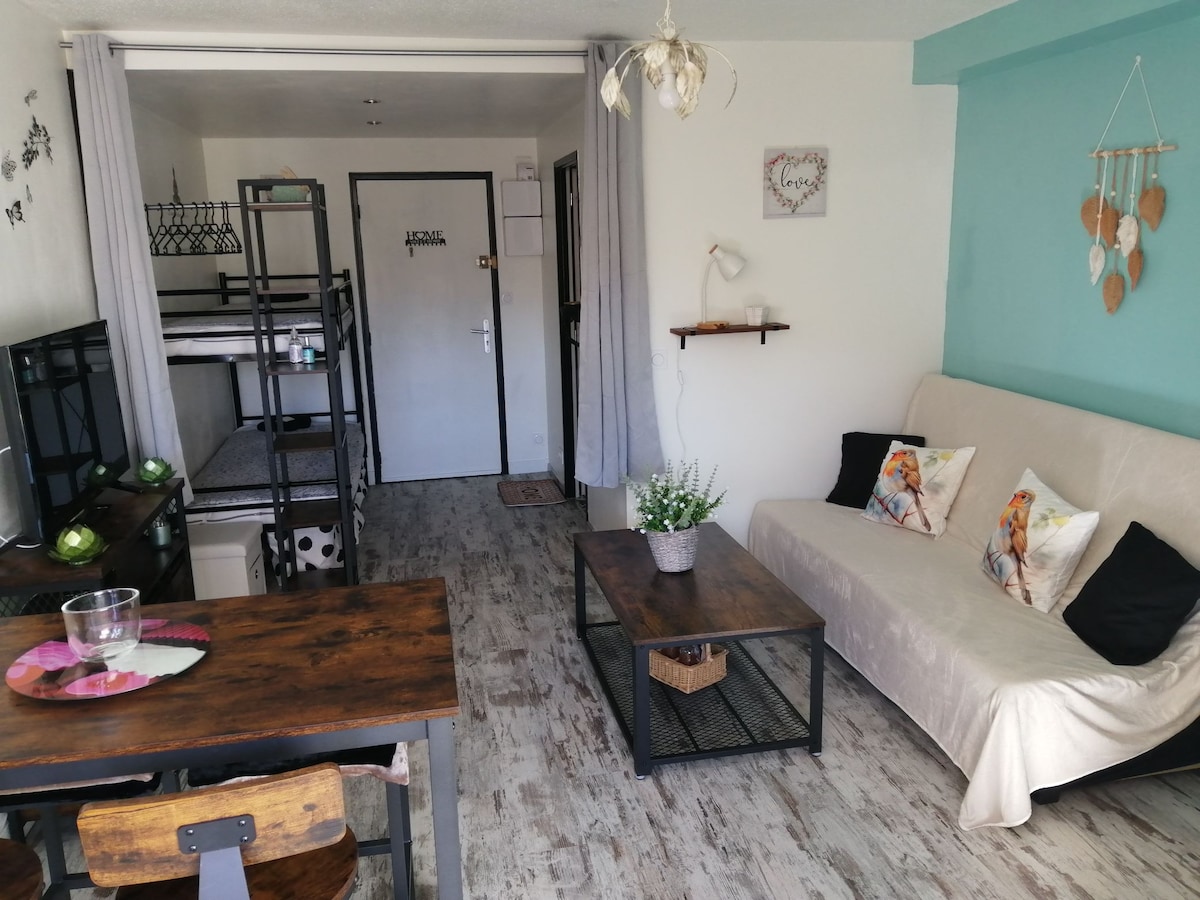 A multifunctional living space features a cozy seating area with a neutral-colored sofa and a wooden coffee table. A dining table with wooden chairs is positioned to one side, while a bunk bed is visible in the background. Bright light enters through a partially visible doorway.