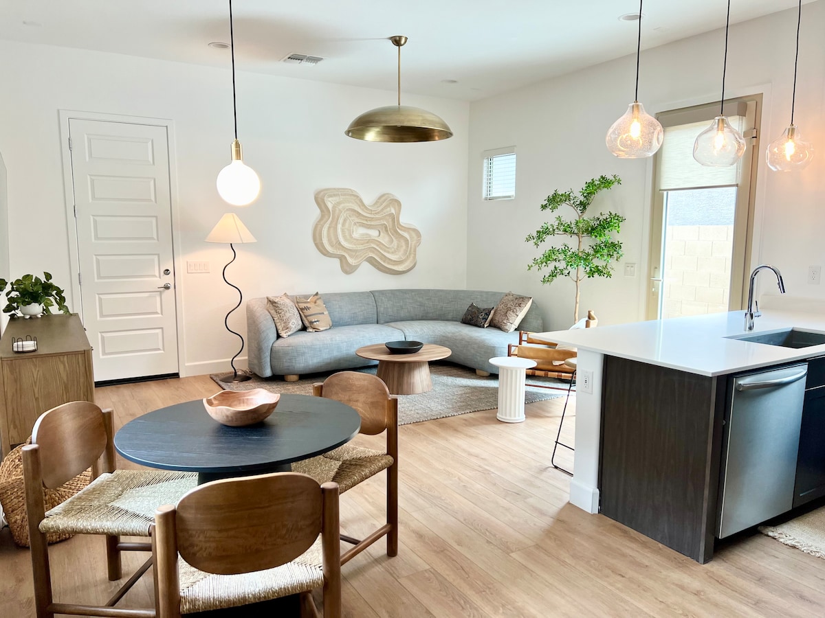 A modern living space features a curved sofa paired with a circular coffee table, enhancing comfort and relaxation. A dining area holds a round table surrounded by four wooden chairs. Natural light illuminates the room through large windows. Stylish pendant lighting adds a contemporary touch.
