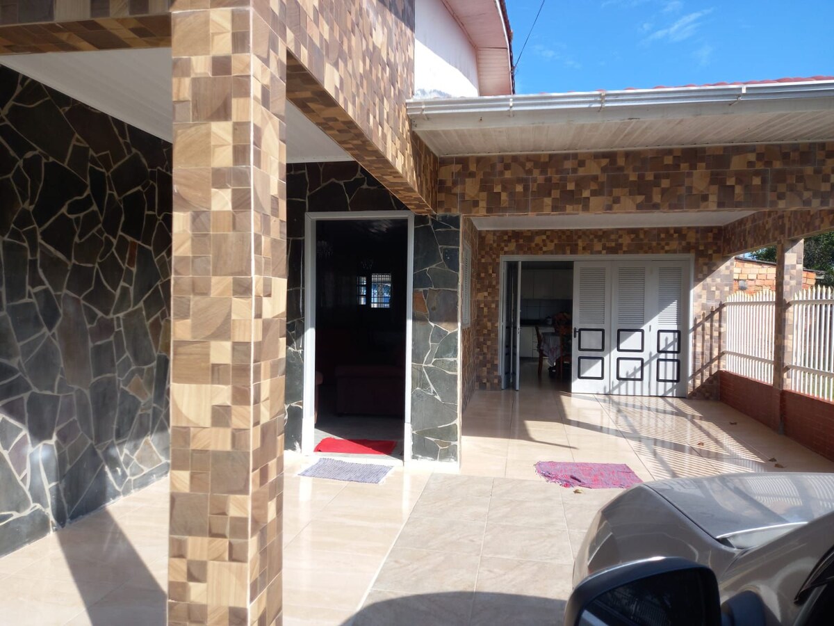 The entrance area of the house features a combination of textured stone and tiled surfaces. A covered porch provides shelter, with a red doormat leading to the main door. Additional spaces for seating are visible, along with an open view towards the outdoors.