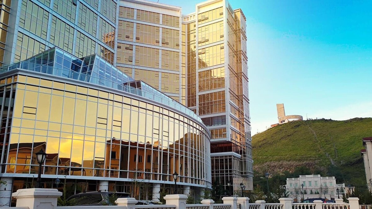 The luxury apartment building is characterized by its modern architecture, featuring a façade of mirrored glass that reflects the surrounding hillside. A nearby monument can be seen on the hill in the distance, adding to the overall appeal of the setting.