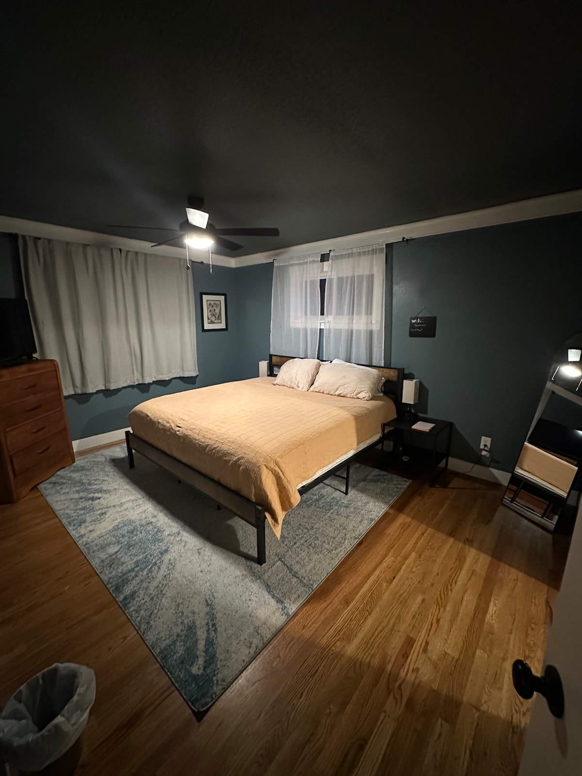 A spacious master bedroom is featured, highlighted by a king bed covered in a soft beige blanket. Light from a ceiling fan and bedside lamps illuminates the room, which includes a wooden dresser and a small nightstand. A large area rug and curtains add subtle texture.