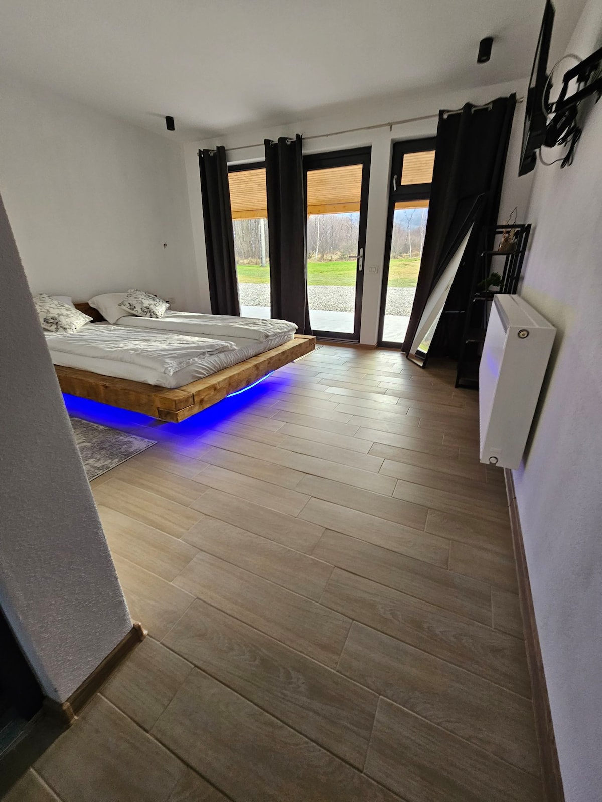 A spacious sleeping area features a wooden bed with integrated lighting along the base. Large glass doors allow natural light to fill the room, while curtains are drawn to the sides. A sleek mirror and modern heating unit are also present, enhancing the overall functionality.