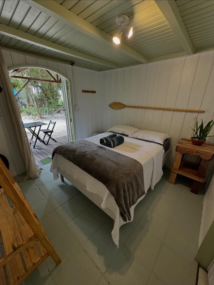 A bright sleeping area features a spacious bed adorned with layered blankets and decorative cushions. Natural light enters through glass doors leading to an outdoor space. Wooden furnishings, including a small table and a rustic side table with a plant, enhance the decor.