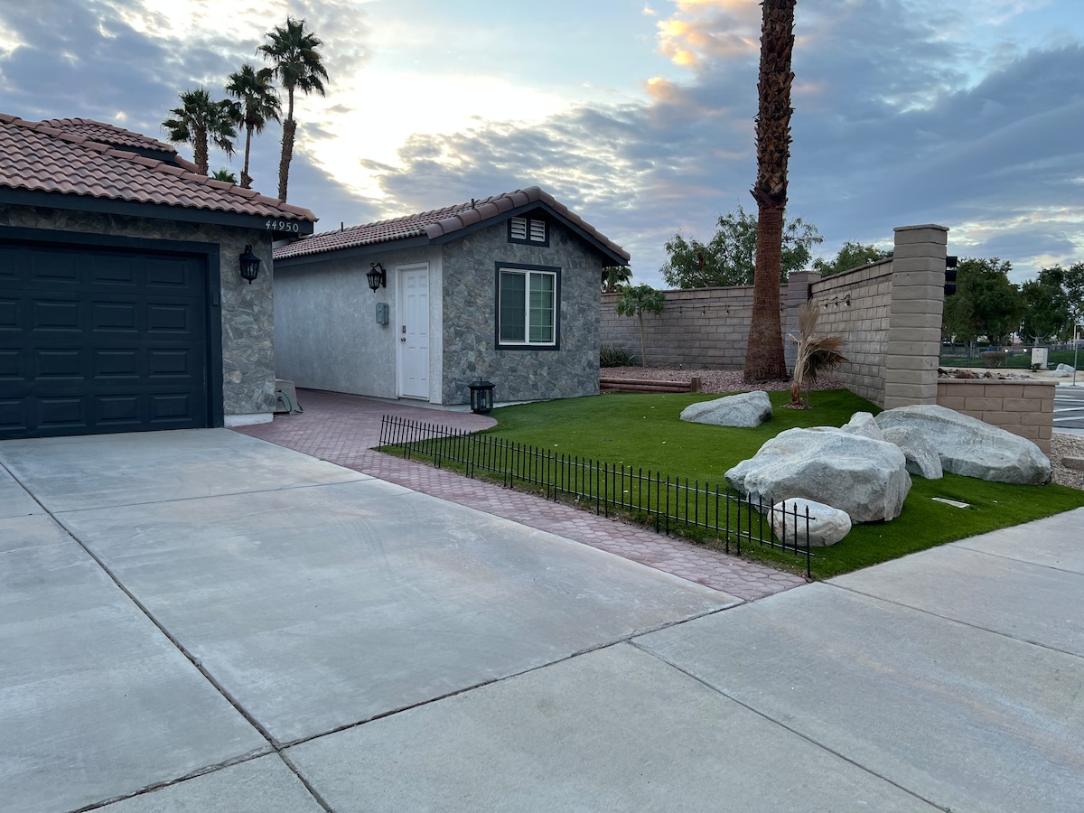 An exterior view of a casita is displayed, featuring a stone façade complemented by a terracotta-tiled roof. Lush green grass borders the area along with several large rocks, while palm trees and a partly cloudy sky are visible in the background.