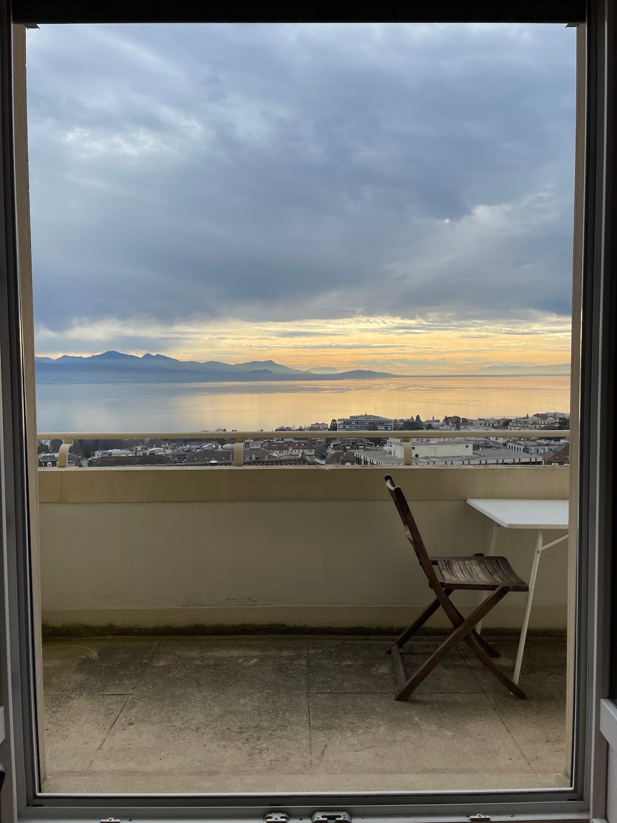 A framed view from a terrace offers a scenic outlook over the Leman valley, showcasing water and distant mountains under a cloudy sky. A small table and a wooden chair are positioned on the terrace, inviting relaxation while enjoying the landscape.