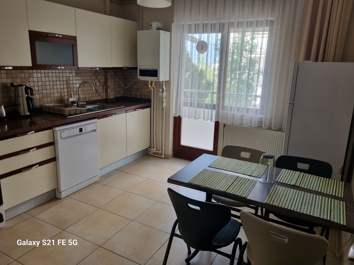 The kitchen area features modern cabinetry and appliances, including a dishwasher and refrigerator. A table with seating for four is set beside a large window, allowing natural light to fill the space. Soft beige tiles cover the floor, creating a clean and functional environment.