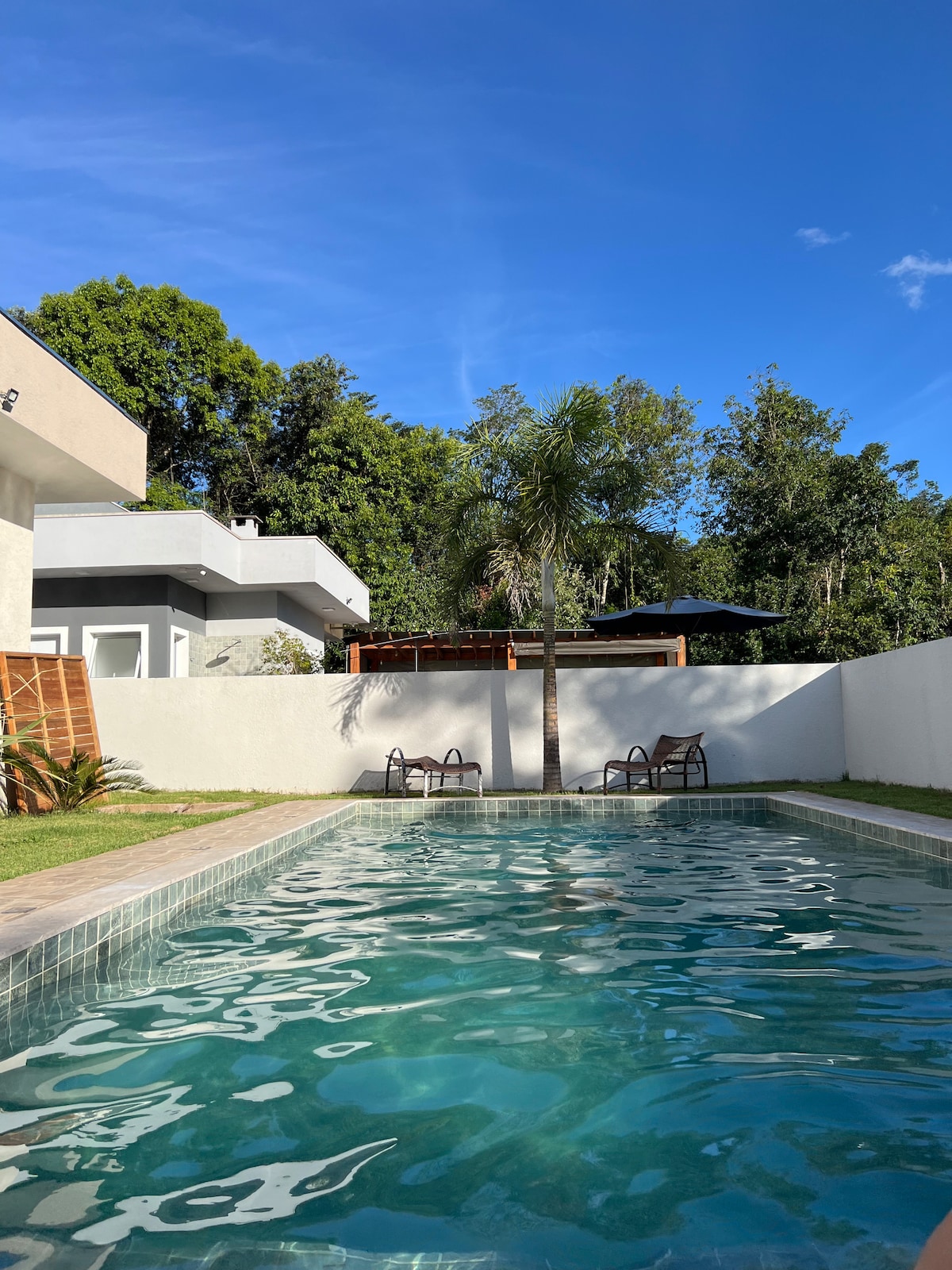 A clear view of the swimming pool is presented, reflecting the blue sky above. Two lounge chairs are positioned on the poolside, surrounded by lush greenery and a white wall, creating a serene outdoor setting.