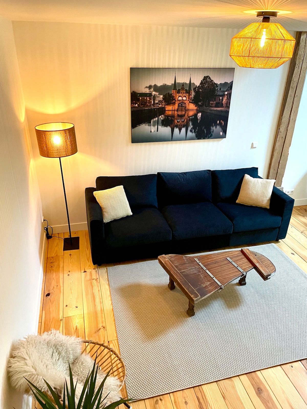 A comfortable living space features a dark blue sofa with two light-colored cushions. A wooden coffee table sits on a neutral area rug. A floor lamp and pendant light provide soft illumination, while a large wall art piece depicts a scenic view of a city at night.
